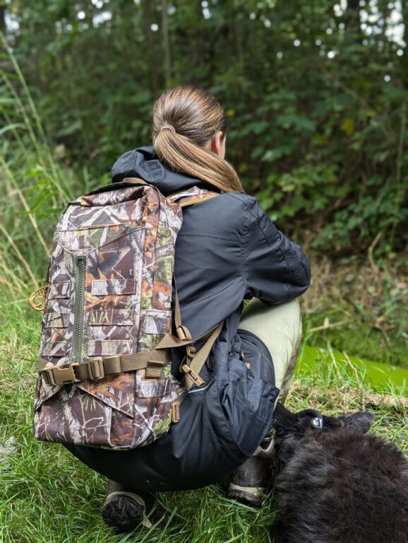 Frau in Outdoor-Kleidung kniet im Wald und trägt einen Tarn-Rucksack, während ein schwarzer Hund neben ihr auf dem Boden ruht.